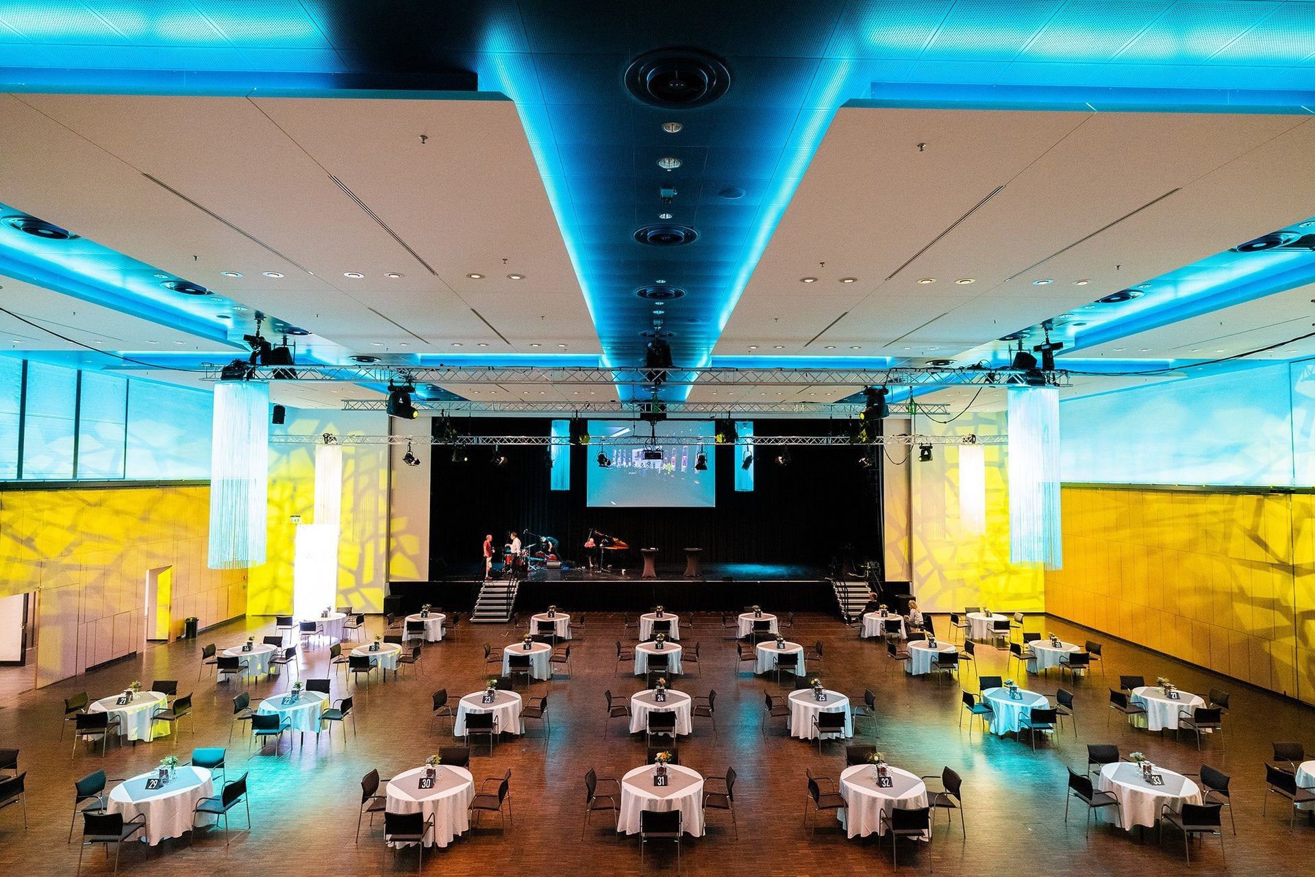Großer Saal des Rhein Sieg Forums mit runden, weiß eingedeckten Tischen auf Parkett, blau leuchtender Decke und Bühne mit Leinwand