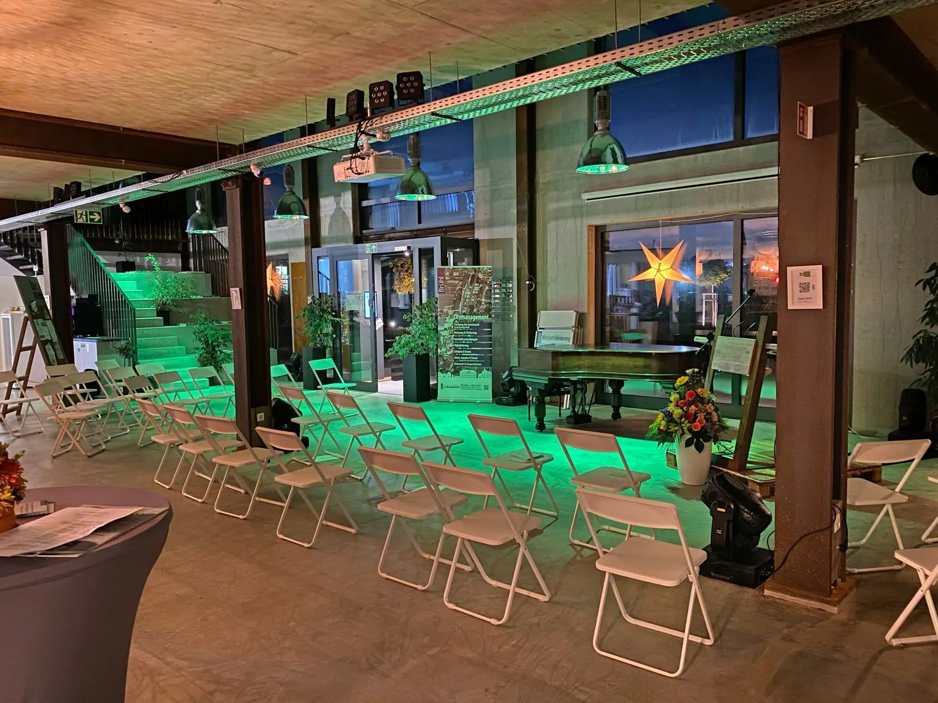 Loft des BRÜNEO in Brühl am Abend mit grün beleuchteter Treppe, Flügel, Pendelleuchten und weißen Stuhlreihen