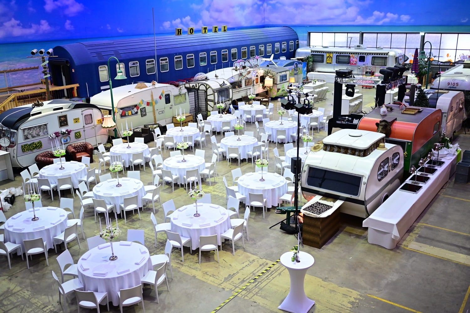 Runde Tische mit weißen Tischdecken und weißen Stühlen in der Halle des BaseCamp Bonn, umgeben von Retro-Wohnwagen und einem blauen Schlafwagen