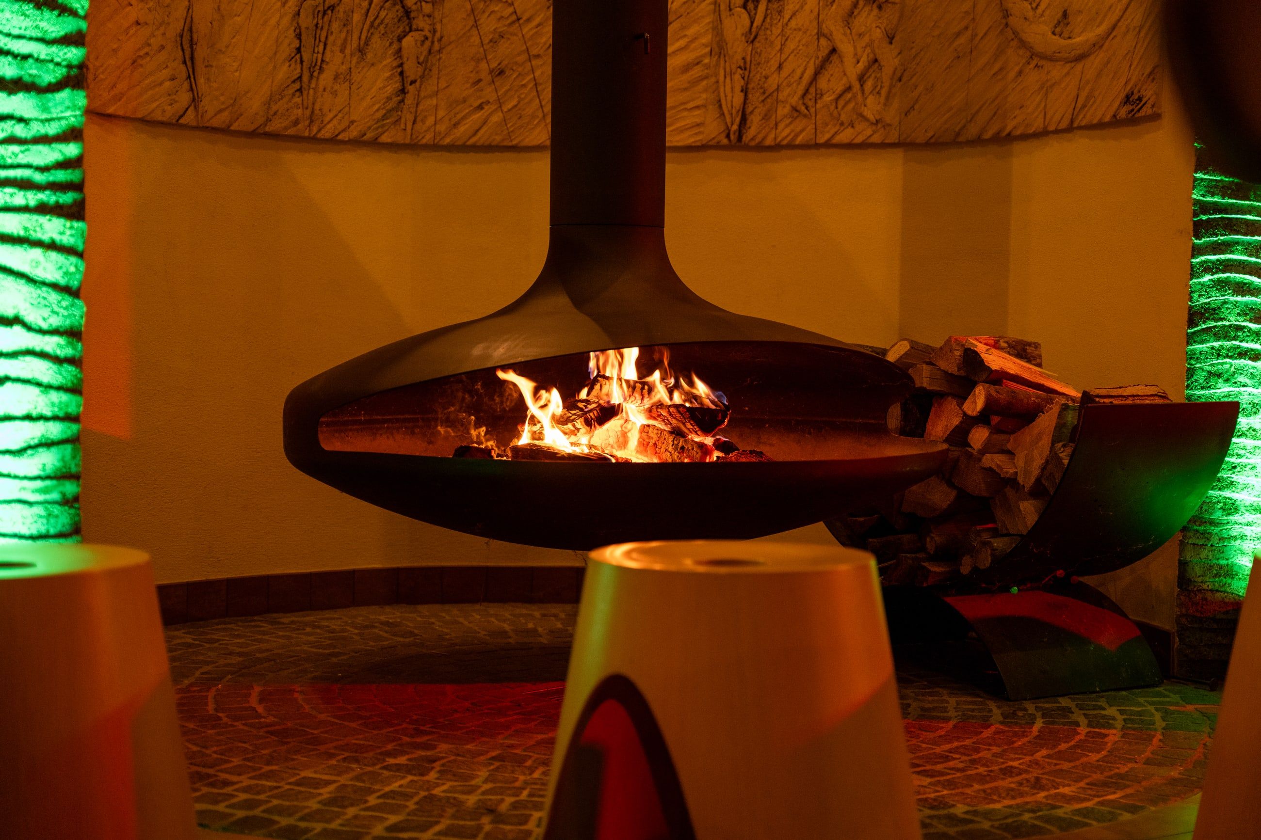 Frei hängender runder Kamin mit offenem Feuer in der Rotunde, davor zwei Hocker und gestapeltes Brennholz