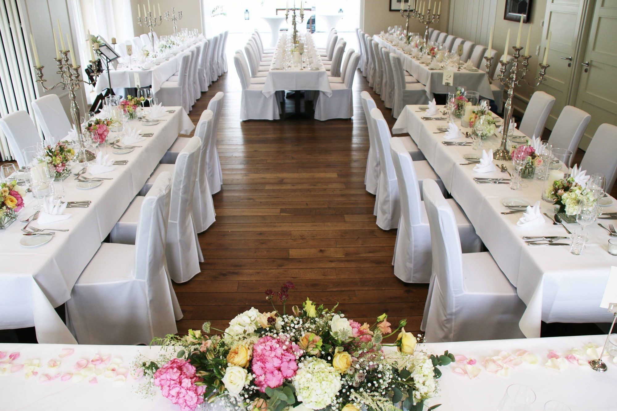 Festsaal des Landhaus Velte mit langen weiß eingedeckten Tafeln, Hussenstühlen und Blumen auf Parkettboden