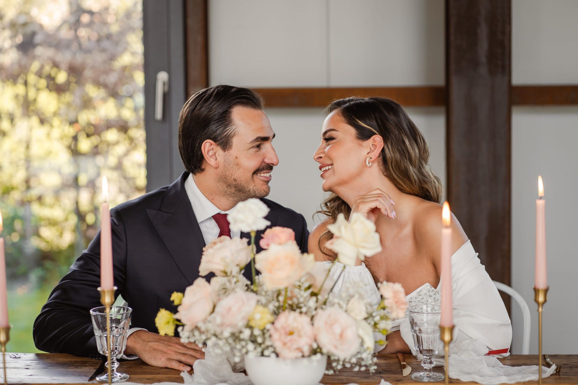 Brautpaar an einer Hochzeitstafel mit Kerzen und Rosen im BRÜNEO, im Hintergrund große Fenster ins Grüne