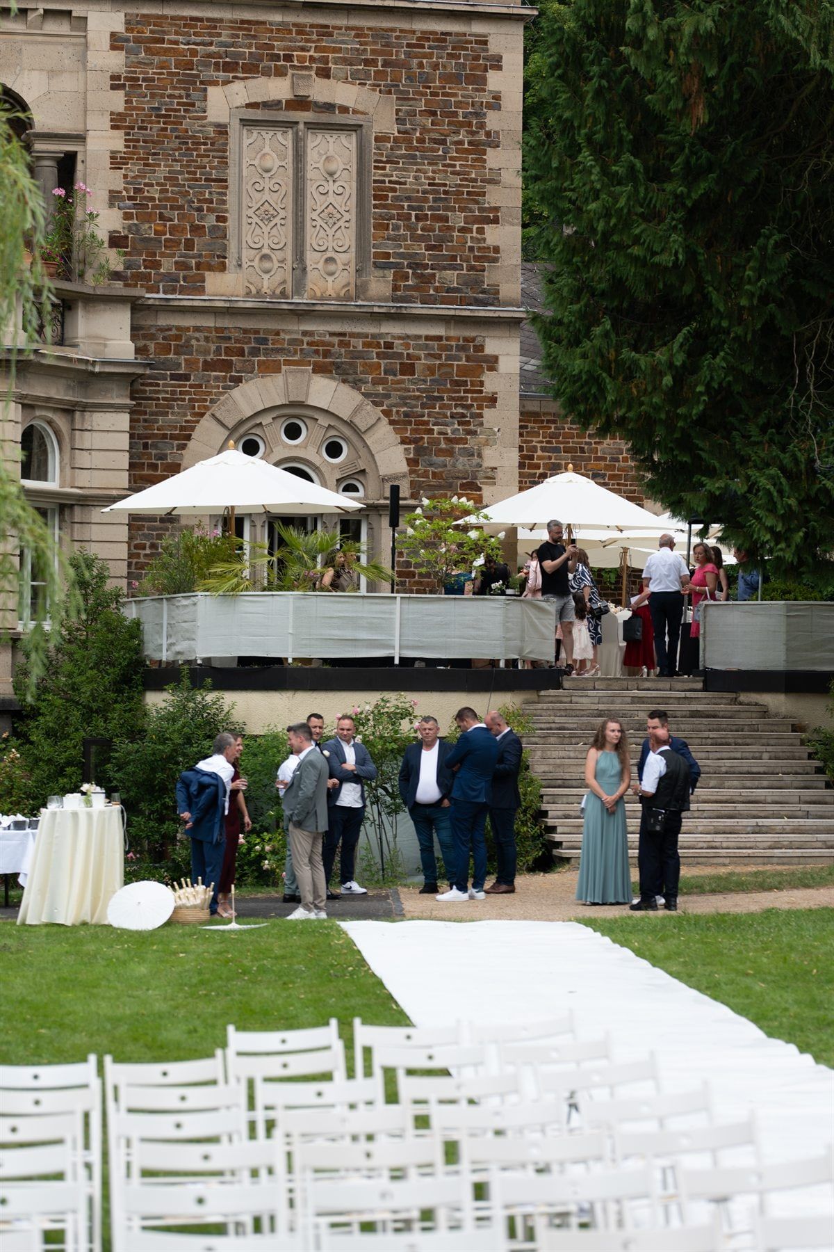 Weiße Stuhlreihen und Teppich für die freie Trauung im Park, dahinter Gäste vor der Terrasse von Schloss Vettelhoven