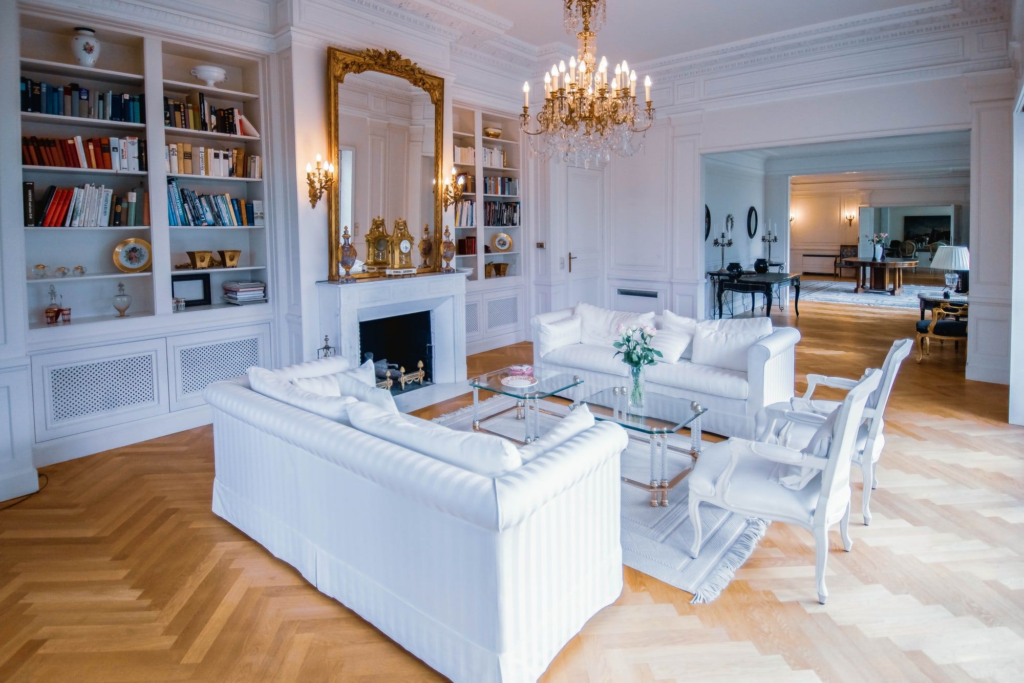Heller Salon der Villa Heckenfels mit Kamin, Bücherwänden, Goldspiegel, Kronleuchter und weißen Sofas auf Fischgrätparkett