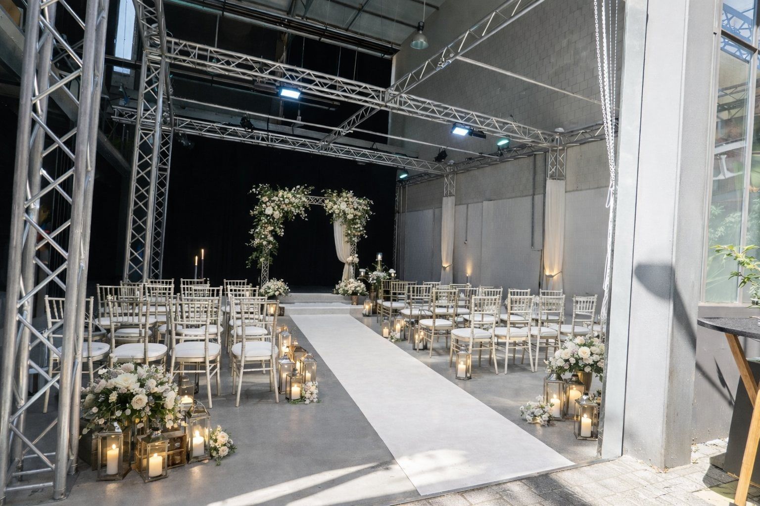 Freie Trauung im Bauwerk Köln, weiße Stühle beidseits eines Gangs unter Traversen, Blumenbogen und Laternen