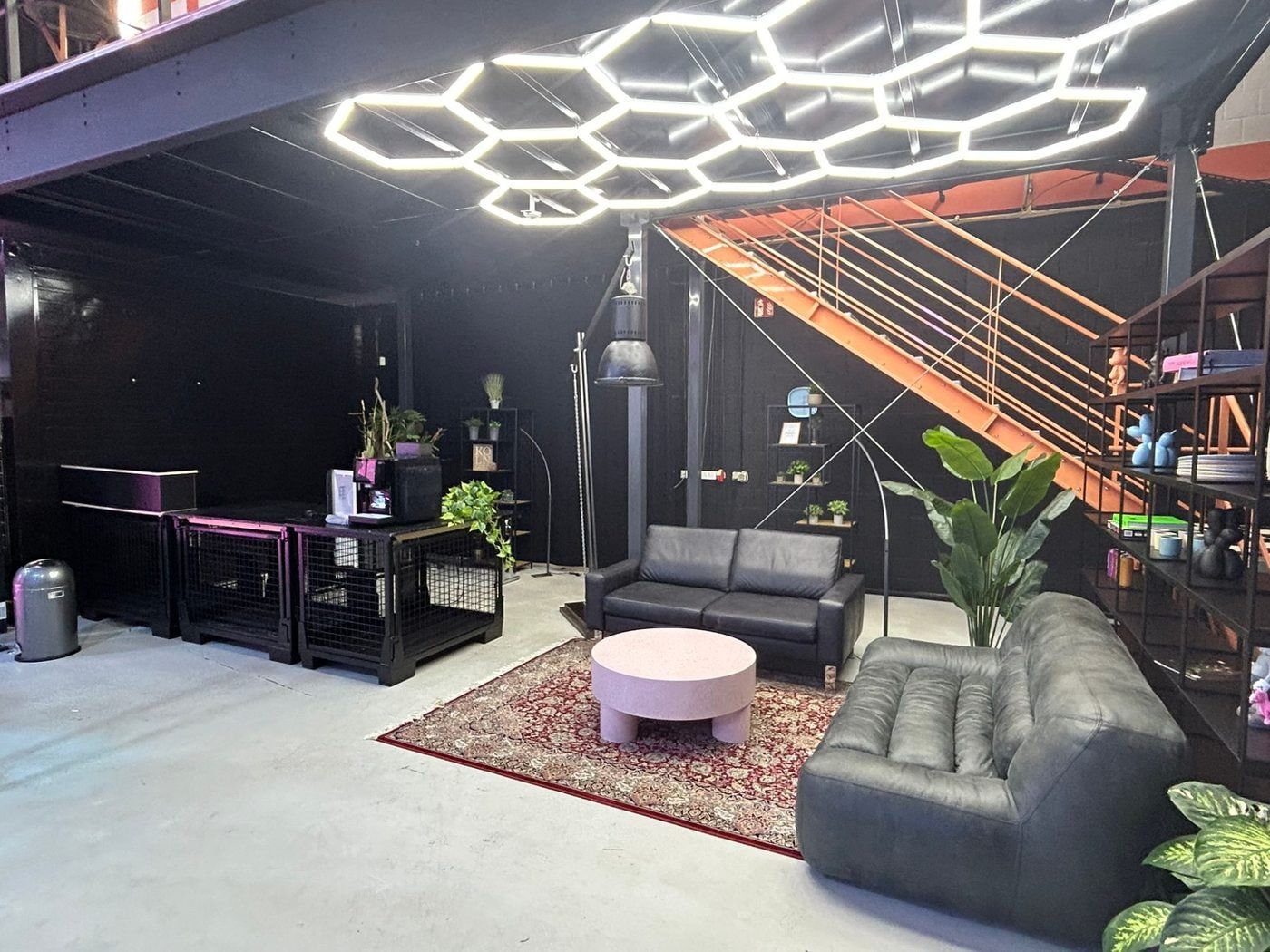 Lounge unter der Galerie des Bauwerk Studios mit Wabenleuchten, dunklen Ledersofas, Theke und orangefarbener Stahltreppe