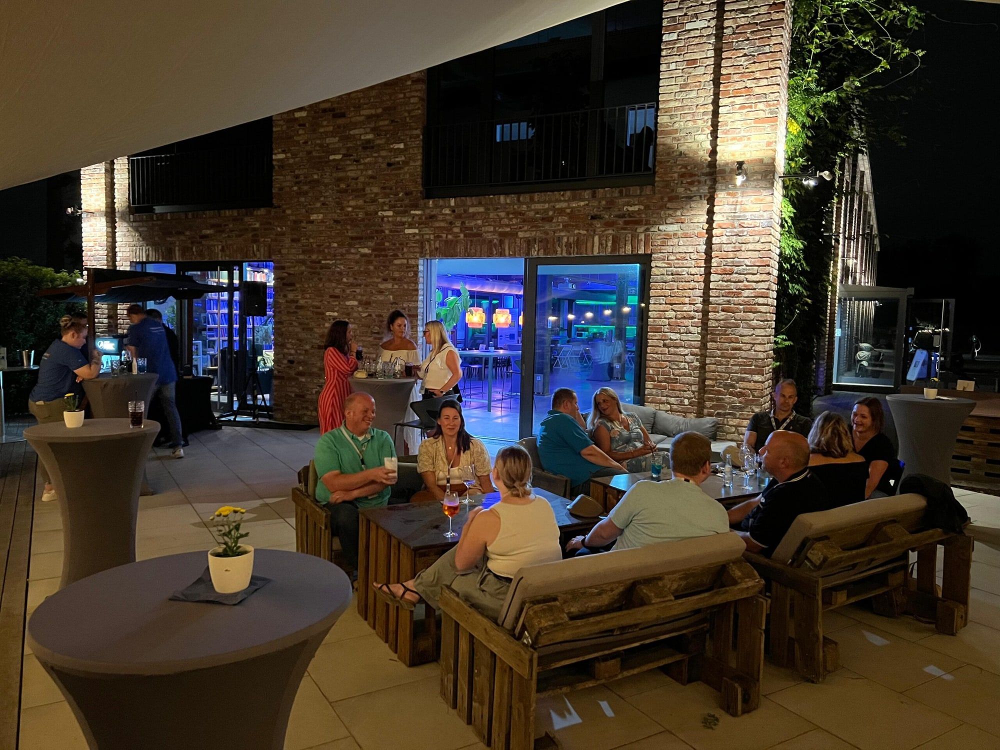 Gäste am Abend auf der Terrasse vor der beleuchteten Klinkerfassade des BRÜNEO in Brühl, Stehtische und Palettenmöbel