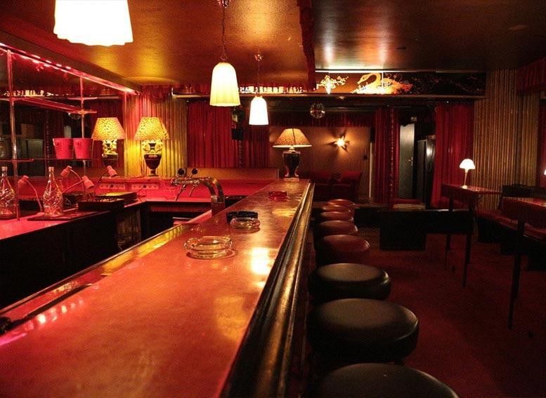 Lange Theke im Untergeschoss der Kokett Bar mit dunklen Barhockern, Tischlampen und roten Vorhängen