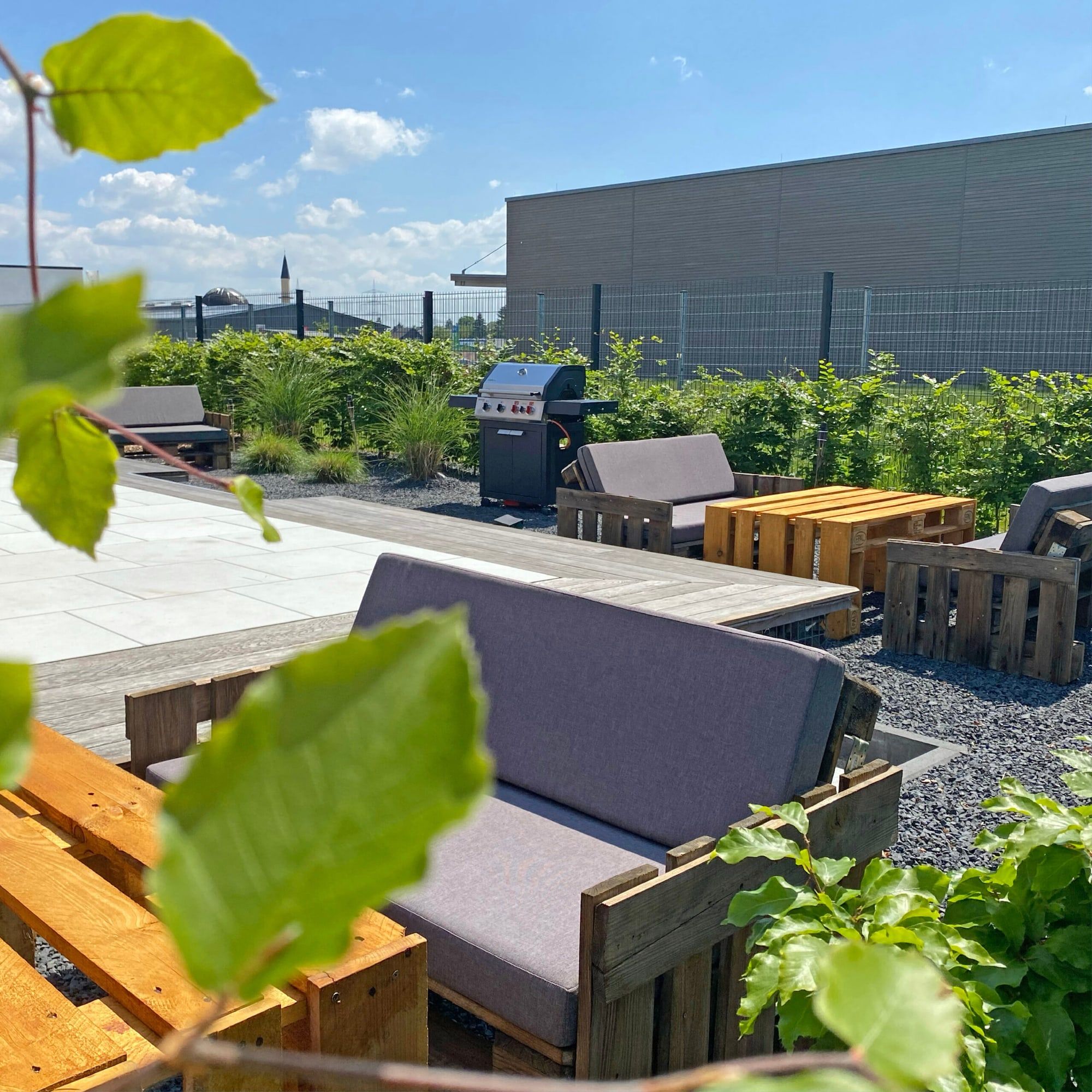 Lounge-Ecke auf der Terrasse des BRÜNEO mit Palettensofas, Holzbänken, Gräsern und blauem Sommerhimmel