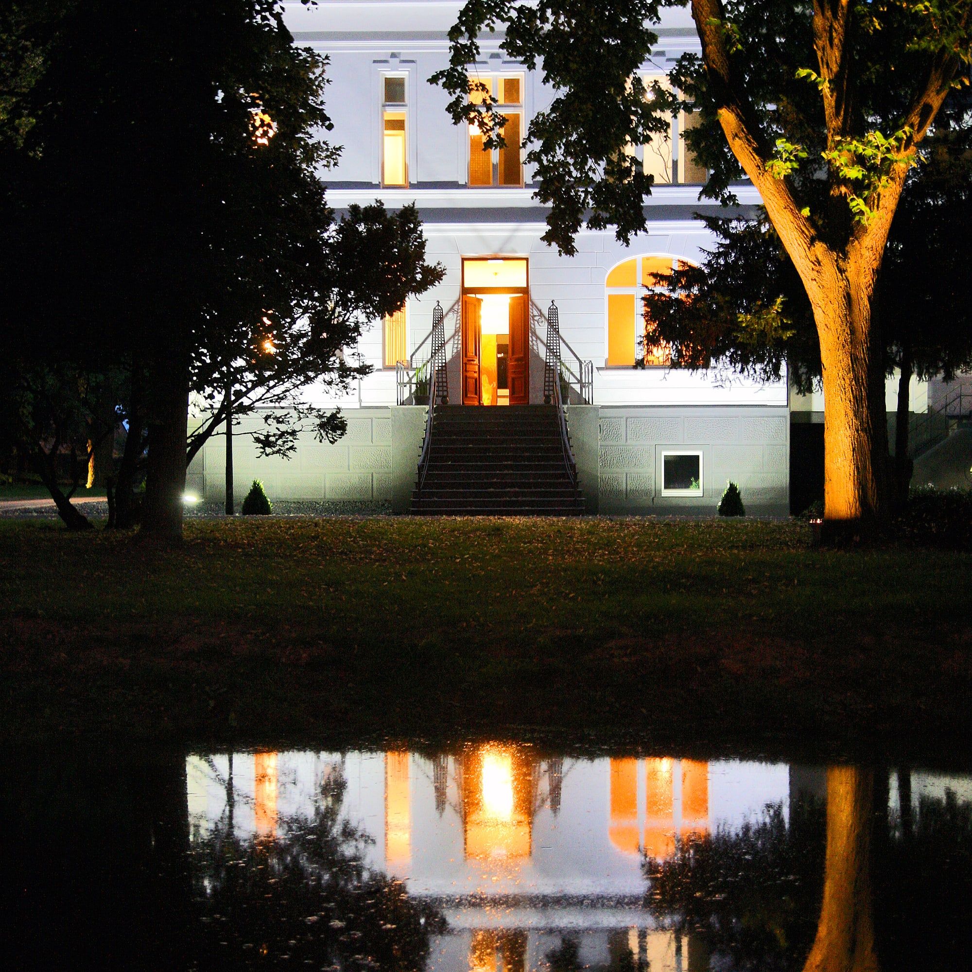 Die beleuchtete Villa des Andreashauses am Abend, Lichter und Bäume spiegeln sich im Teich des Parks