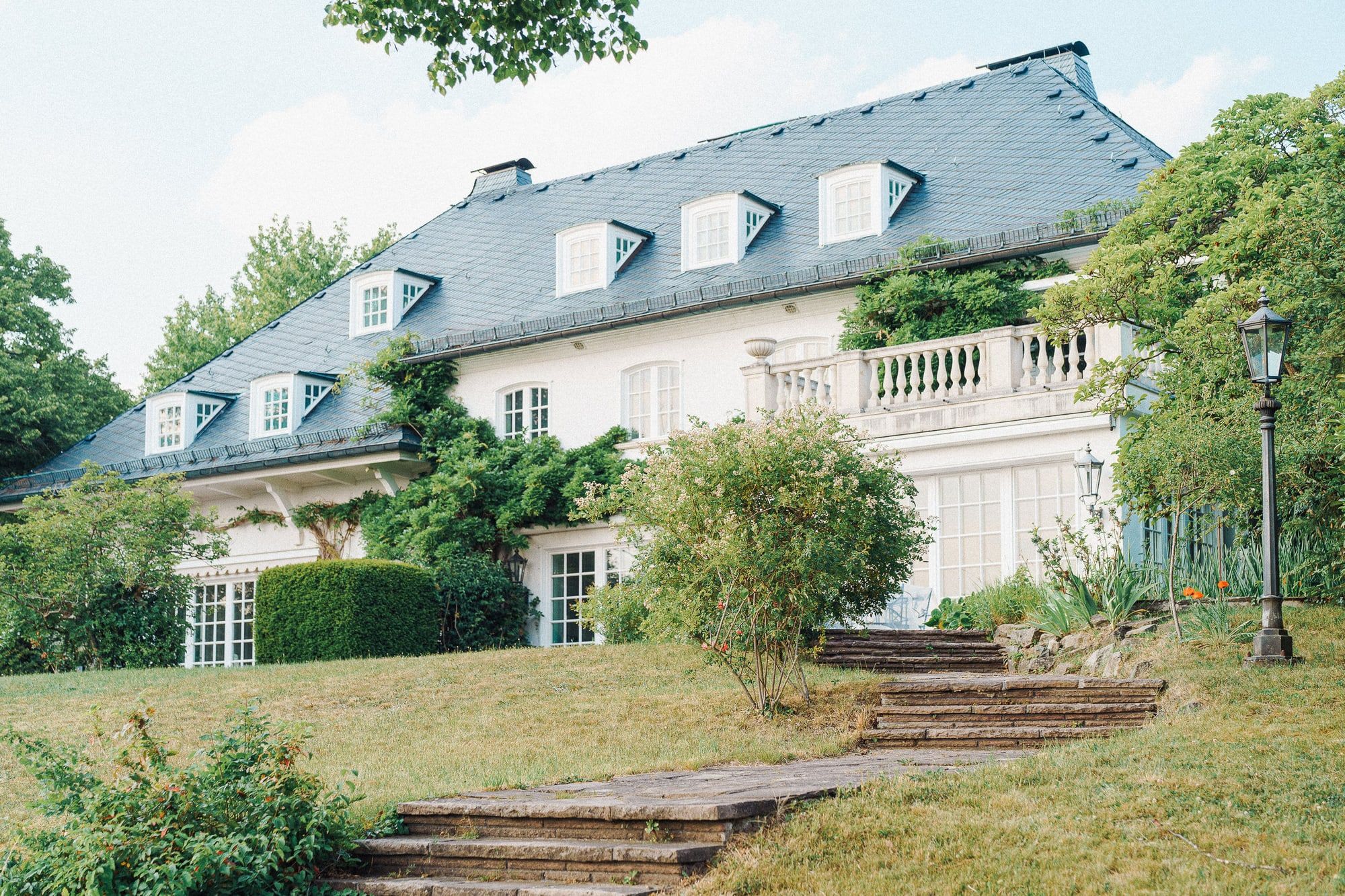 Die Villa Heckenfels vom Garten aus gesehen, weißes Herrenhaus mit Schieferdach, Terrasse und Steinstufen im Rasen