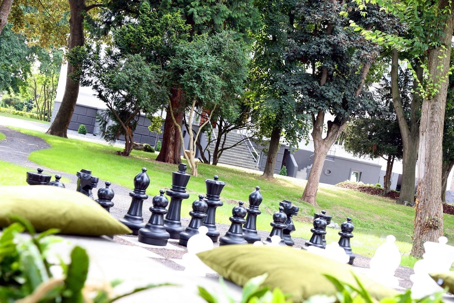 Park des Andreashauses mit großem Gartenschach, Sitzkissen und altem Baumbestand an einem Sommertag