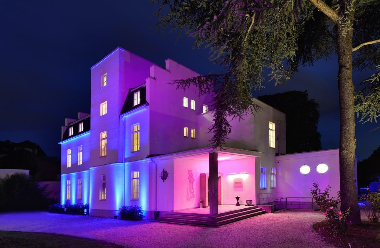Eingang der Villa Leonhart bei Nacht, die weiße Fassade in Violett und Blau angestrahlt, davor eine alte Zeder