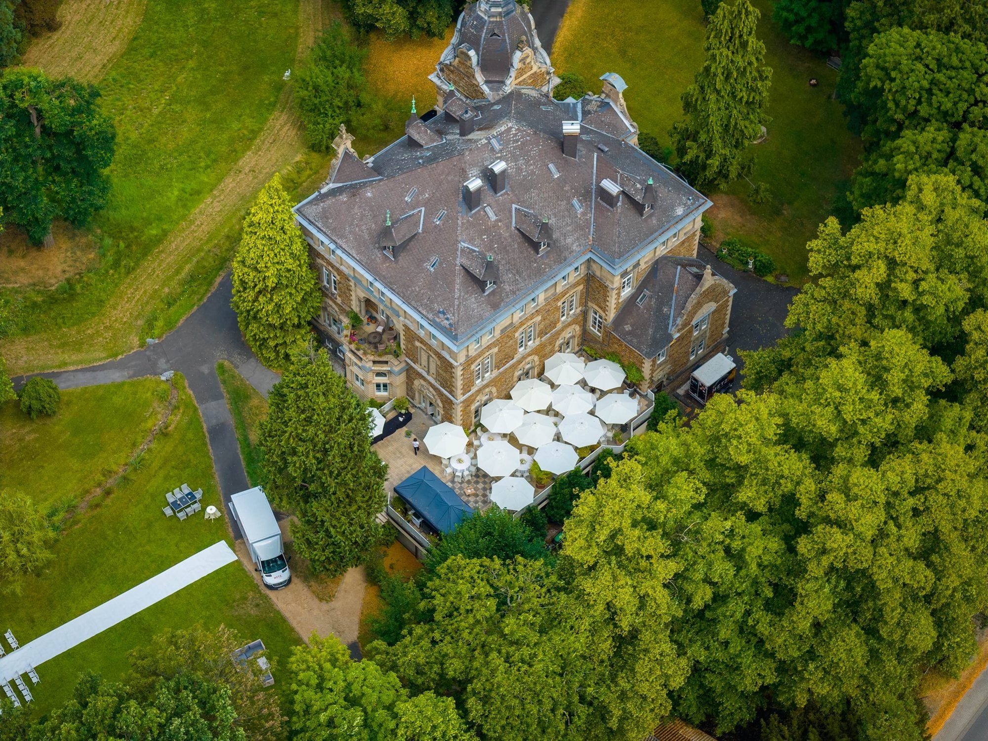 Schloss Vettelhoven von oben mit Schieferdach, Terrasse voller Sonnenschirme und dem Park rundherum