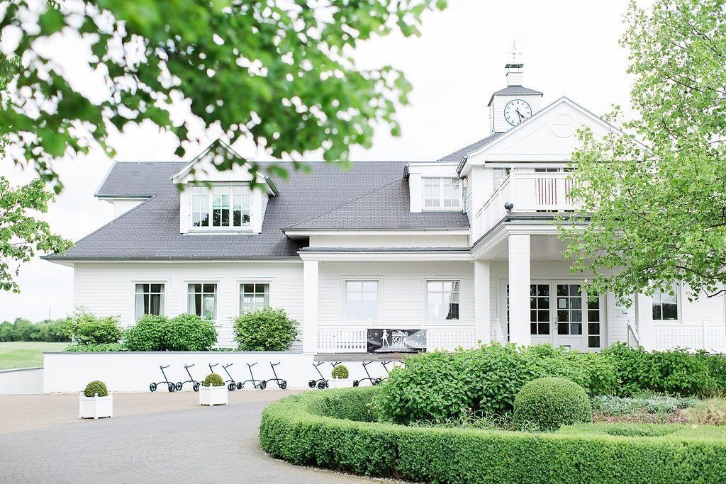 Vorderseite des Landhaus Velte mit Uhrtürmchen, rundem Buchsbeet und Bäumen an einem hellen Tag